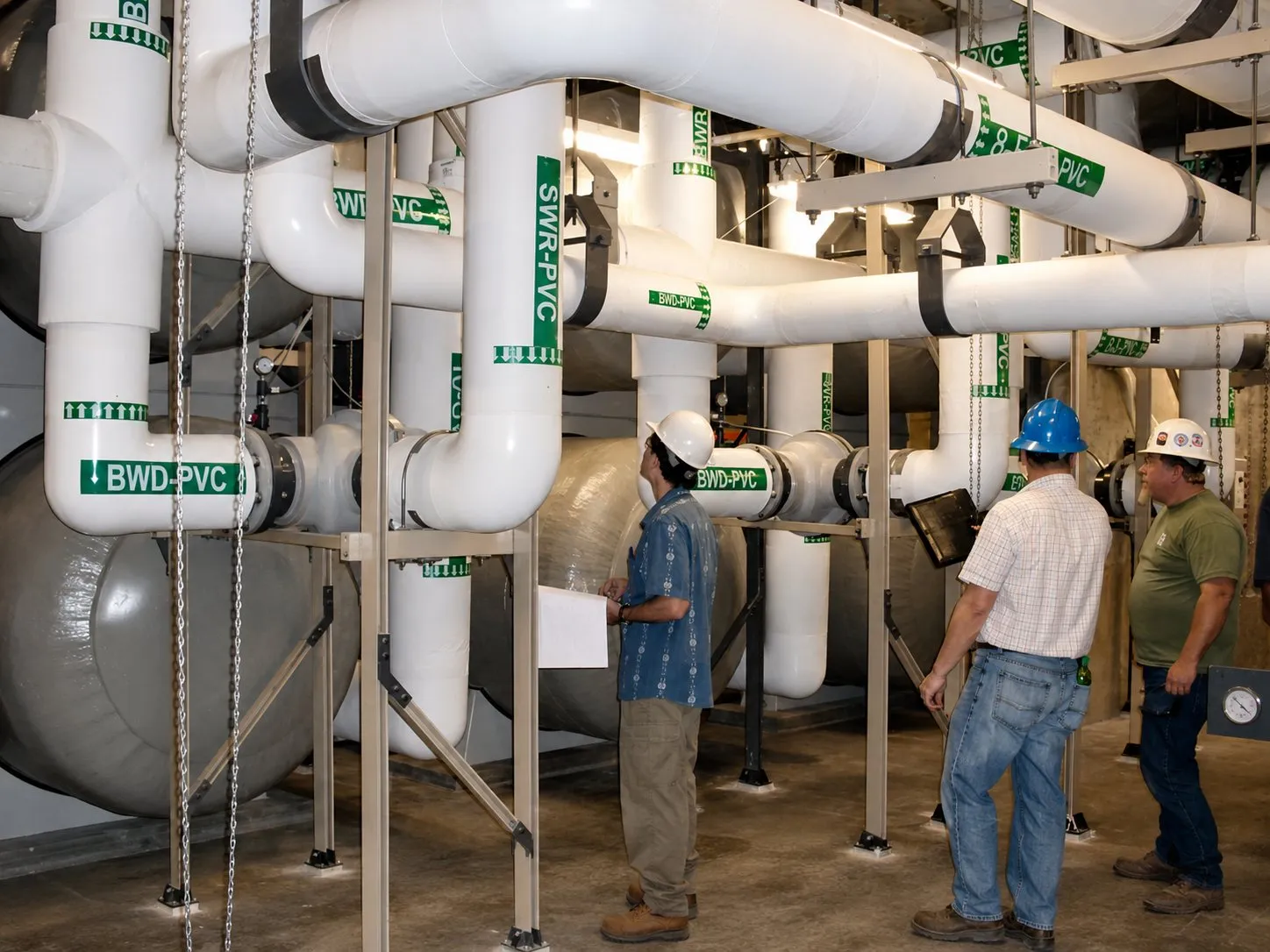 Three staff in hard hats reviewing drawings beneath labeled process piping in a life support system gallery